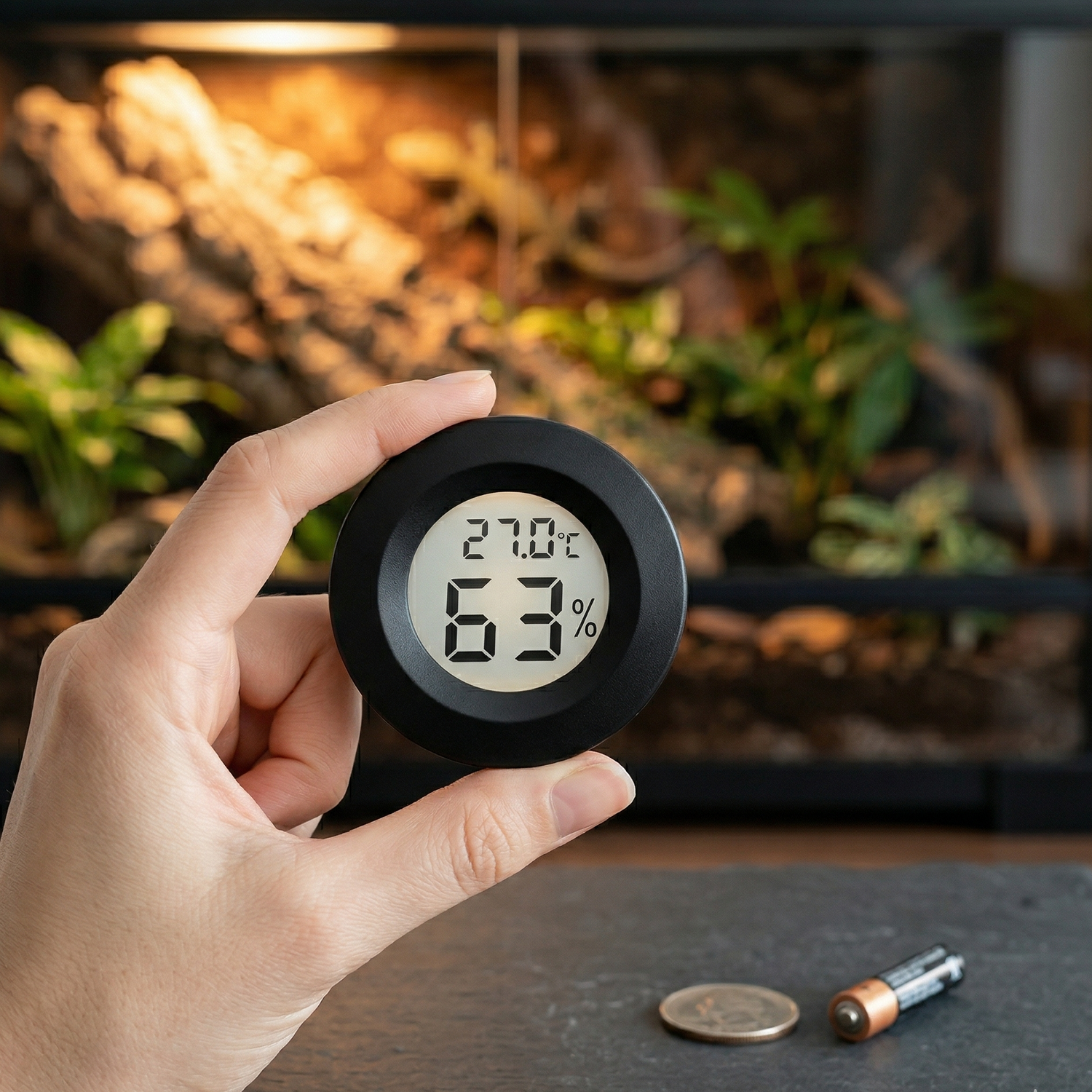 Main élégante tenant hygromètre thermomètre LCD noir compact 45mm 
entre pouce index montrant taille miniature, écran 27.0°C 63% net 
lisible, pièce monnaie batterie AAA surface ardoise échelle référence, 
terrarium décoré bokeh chaud ambre gecko silhouette arrière-plan flou, 
lumière naturelle froide premier plan contraste chaud derrière, 
format JPG 1:1