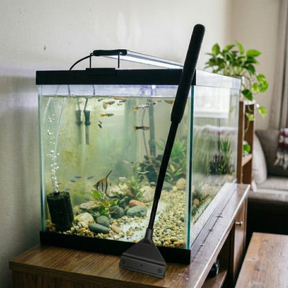Nettoyeur vitre noir (exact référence design) posé leaning against front glass aquarium ou resting rim montrant full length casually tool not being used positioned naturally just finished cleaning, aquarium maison normal 60-80cm wooden stand TV cabinet eau normale natural slight haze not perfectly clear poissons réguliers guppies tetras goldfish common pet store fish basic gravel plastic plant simple decorations lived-in typical family aquarium not elaborate aquascape, background REGULAR living room home sp