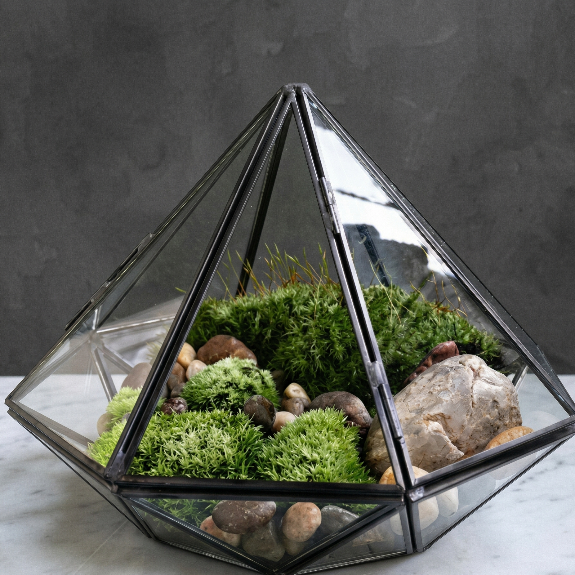Terrarium géométrique diamant verre clair cadre métal noir mat précis, 
facettes géométriques multiples, intérieur mousse verte vibrante 
galets rivière pierre quartz cristal bois flotté, fond blanc marbre 
poli éclairage joaillerie qualité, facettes verre lumière différente 
profondeur, format JPG 1:1